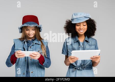 Fröhliche interracial Kinder in modischen Denim-Kleidung und Hüte mit Smartphone und digitales Tablet isoliert auf grau Stockfoto