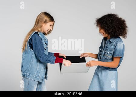 Lachend interracial Kinder in Denim Kleidung, die Laptop von jedem Andere isoliert auf grau Stockfoto