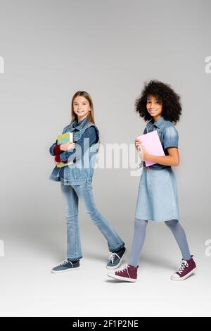 Trendy interracial Schülerinnen in Denim Kleidung und Gummischuhe zu Fuß mit Lehrbücher auf grau Stockfoto