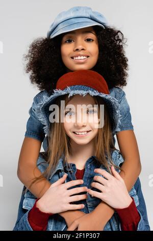 Happy interracial Kinder in modischer Kleidung und Hüte umarmen und Blick auf die Kamera isoliert auf grau Stockfoto