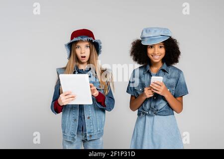 Aufgeregt interracial Kinder in trendigen Kleidung mit Smartphone und digital Tablett isoliert auf grau Stockfoto