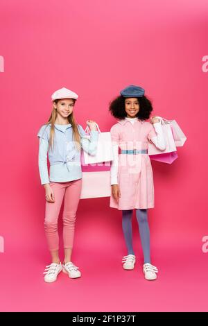 In voller Länge Ansicht von interracial Kinder im trendigen Frühjahr Outfit Posiert mit Einkaufstaschen auf rosa Stockfoto