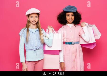 Fröhliche interracial Kinder in modischer Kleidung posiert mit Einkaufstaschen Auf Rosa Stockfoto