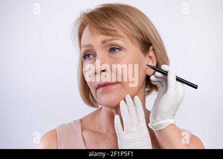 Arzt Zeichnung Marken auf ältere Frau Gesicht für die kosmetische Operation Operation auf hellen Studio Hintergrund. Reife Dame immer bereit für aestetic operatio Stockfoto