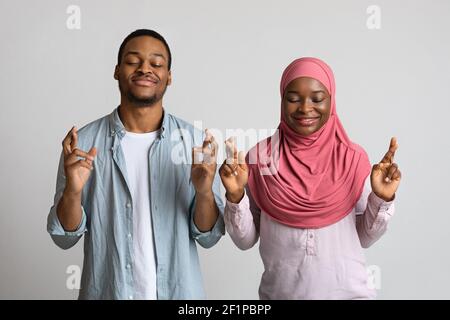 afroamerikanisch-muslimisches Paar mit geschlossenen Augen und gekreuzten Daumen über grauem Studiohintergrund stehend. Abergläubischer schwarzer Mann und Frau in Hijab p Stockfoto