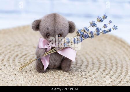 Ein niedlicher Teddybär mit rosa Schleife sitzt auf einem Weidenhintergrund und hält einen Strauß getrockneter Lavendelblüten. Stockfoto
