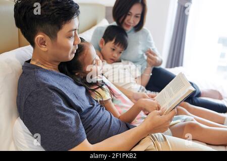 Vietnamesischer Mann liest Interessantes Buch für seine Kinder und seine Frau Sie liegen alle auf dem Bett Stockfoto