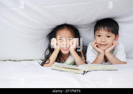 Glücklicher kleiner Bruder und Schwester, die unter der Decke mit geöffnet ruhen Buch und Blick auf die Kamera Stockfoto