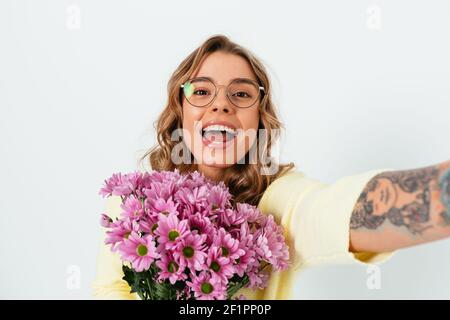 Schöne lächelnde junge Frau trägt Brille hält Bouquet von frischen Blumen macht Selfie, während auf weißem Hintergrund stehen. Stockfoto