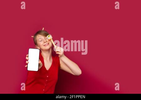 Zarte junge blonde Frau in roten T-Shirt zeigt Mock-up, weißen Bildschirm Handy , hält Philadelphia Sushi. Traditionelle japanische Küche Stockfoto