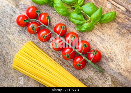 Reife Tomaten umgeben von frischen Basilikumblättern und getrockneten Spaghetti Auf einem Holzbrett Stockfoto