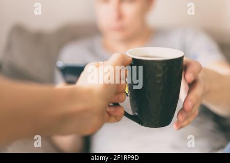Zugeschnittenes Bild eines Mannes mit Smartphone, der eine Tasse heißen Tee oder Kaffee aus der Hand einer Frau nimmt, Nahaufnahme Stockfoto