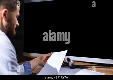 Zugeschnittenes Foto eines Mannes mit Papieren, der vor einem schwarzen Monitor sitzt. Schwarzer Bildschirm mit Platz für Text oder Werbung. Stockfoto