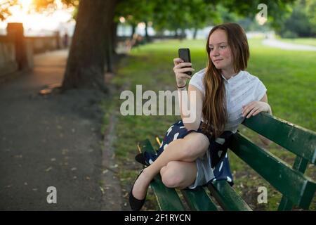 Junges Mädchen sitzt mit einem Smartphone auf einer Bank in einem Park. Stockfoto