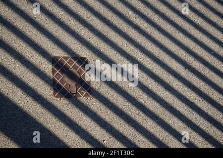 Eiserne Zugangsabdeckung auf einer Straße. Metallabdeckung auf einer Straße. Zaunschatten auf Asphalt. Abstrakter Hintergrund. Stockfoto