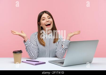 Positiv lächelnde Frau Büroangestellte in gestreiften Hemd zuckenden Schultern und werfen ihre Hände oben sitzen am Arbeitsplatz, weiß nicht Antwort. Stu Stockfoto