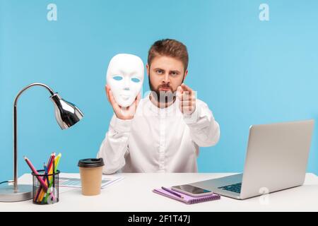 Serious bärtigen Mann Büroarbeiter hält weiße Gesichtsmaske und zeigt Finger auf die Kamera, beschuldigen Sie für das Verstecken Identität, Doppelzüngigkeit. Studio-Sho Stockfoto