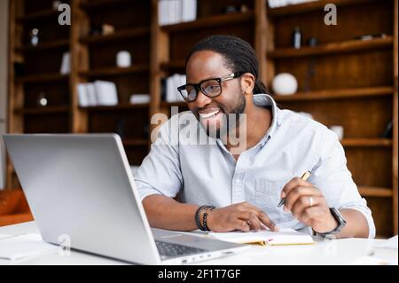 Lächelnder afroamerikanischer Kerl nimmt Notizen, die Webinare auf dem Laptop anschauen, Geschäftsaufgaben erforschen, studiert ein männlicher Student online, hört Videolectures, schreibt auf, E-Learning-Konzept Stockfoto