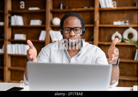 Der afroamerikanische Mann ist Callcenter-Mitarbeiter oder Support-Kundenbetreker. Ein intelligenter dunkelhäutiger Mann in einer Brille verwendet ein schnurloses Headset und einen Laptop, um an seinem Arbeitsplatz online zu sprechen Stockfoto