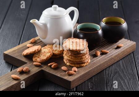 Schwarzer Tee in chinesischen schwarzen Schüsseln serviert mit Cookies und Mandeln auf dunklem Eichenbrett auf schwarzem verbranntem Holzhintergrund Stockfoto