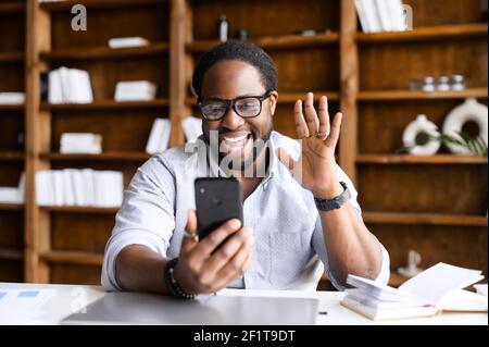 Überglücklich afroamerikanischen Kerl mit einem Smartphone für Videoanruf sitzen im Büro, ein Mixed-Rennen fröhlich jungen Mann winkt hallo, froh, Gesprächspartner, Video-Meeting, virtuelle Konferenz-Konzept zu treffen Stockfoto