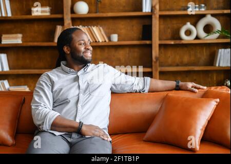 Ein unbeschwerter und fröhlicher afroamerikanischer Kerl ruht auf dem Sofa im stilvollen Wohnzimmer, Bücherregale im Hintergrund, ein lächelnder multirassischer entspannter Mann mit Dreadlocks schaut weg Stockfoto