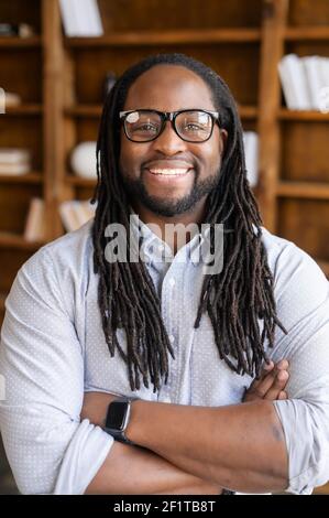 Vertikales Nahaufnahme-Porträt eines fröhlichen afroamerikanischen Geschäftsmannes in stylischen Brillen mit Dreadlocks, ein Kerl steht mit gekreuzten Armen und blickt mit einem freundlichen Lächeln auf die Kamera Stockfoto