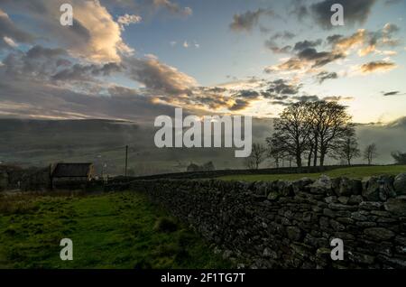 Eine alte Steinscheune in einem Hochland-Ackerland in den North Pennines, mit einer Steinmauer, die darauf hinläuft und sonnenbeschienenen Wolken dahinter Stockfoto