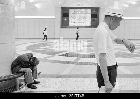 Mann, der die Zeit überprüft und Obdachloser, der auf der Bank am Bahnhof in Tokio, Japan, schläft. Stockfoto