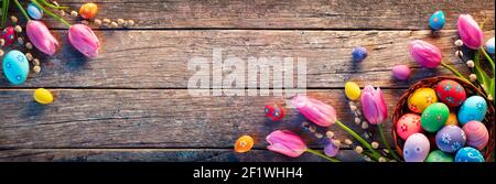 Osterdekoration - Painted Eggs In Vintage Basket On Rustikal Tisch Mit Weidenzweigen Und Tulpen Stockfoto