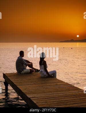 Kreta Griechenland, junge romantische Paar in der Liebe sitzt und umarmt auf hölzernen Pier am Strand in der Sonnenaufgangszeit mit goldenem Himmel. Stockfoto