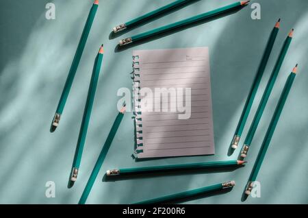 Leeres Blatt Papier und grüne Stifte auf hellblauem Hintergrund mit Schatten. Eine Seite wird aus einem federbelasteten Notizbuch gerissen, umgeben von einem Holzstift Stockfoto