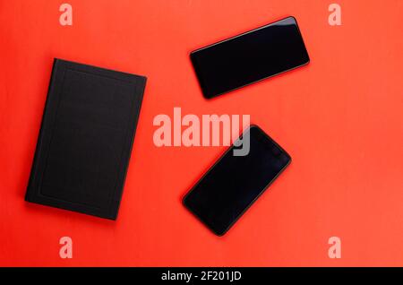 Zwei schwarze Telefone und ein geschlossenes Papierbuch auf rotem Hintergrund. Die Vorteile des Buchlesens Konzept. Flach liegend Stockfoto