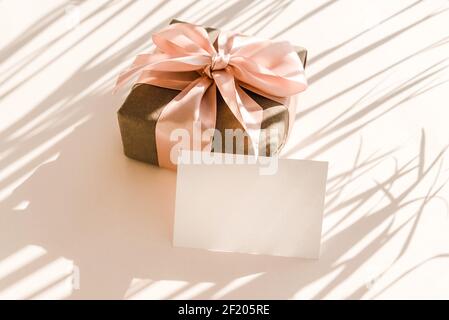 Unbeschriftete Karte und Geschenkbox auf beigem Hintergrund mit schattenförmigen Palmblättern. Minimalkonzept. Stockfoto