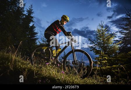 Lächelnder junger Mann im Sportfahranzug Fahrrad bergab mit schönem blauen Himmel auf Hintergrund fahren. Männlicher Fahrradfahrer mit Schutzhelm und Brille, während er nachts den grasbewachsenen Hügel hinunterradelt. Stockfoto