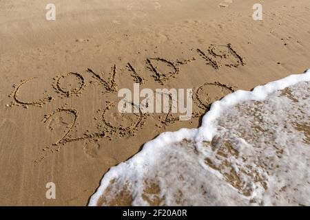 Blick auf Covid-19 und 2020 Text im Sand am Strand Wird von Meereswellen bedeckt und verschwindet Stockfoto