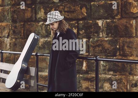 Person in dunklem Mantel und gemustertem Hut steht neben einer Bank mit einem Gitarrenkoffer an einer Steinwand, Farbfoto. Stockfoto