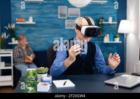 Glücklicher Rentner, der mit einem Headset die virtuelle Realität erlebt. Älterer Mann Rentner im Wohnzimmer sitzen am Schreibtisch mit futuristischen Brille für erweiterte Cyberspace Frau Fernsehen. Stockfoto