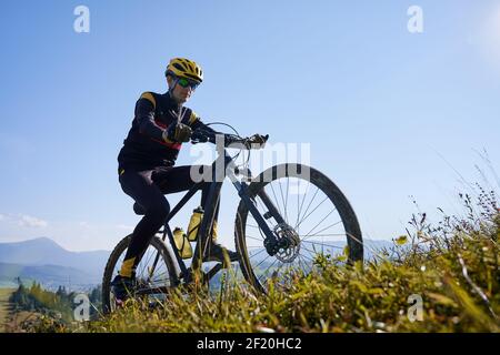 Mann in Schutzhelm und Brille beim Bergfahren am sonnigen Tag mit blauem Himmel im Hintergrund. Männliche Radfahrer im Radanzug bergauf auf auf dem Mountainbike klettern. Konzept von Sport und aktiver Freizeit. Stockfoto