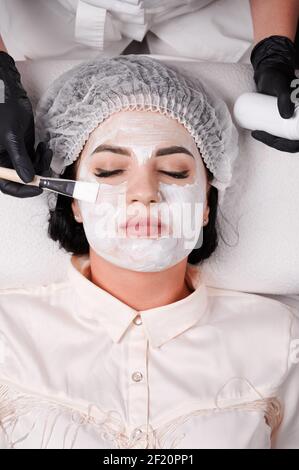 Nahaufnahme der kosmetologischen Hände in schwarzen sterilen Handschuhen Anwendung Reinigung und feuchtigkeitsspendende Maske auf weibliche Client Gesicht. Frau in Einweg-Duschhut erhalten Spa Hautpflege Behandlung im Schönheitssalon Stockfoto