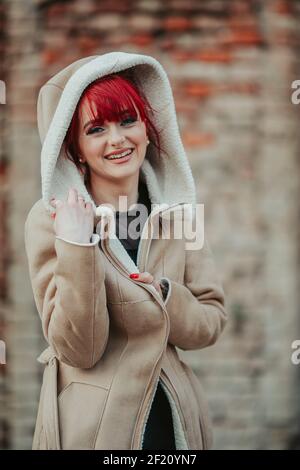 Eine junge schöne lächelnde rothaarige Frau mit Pony trägt ein Warmer Wintermantel mit Kapuze in verschwommenem Hintergrund Stockfoto