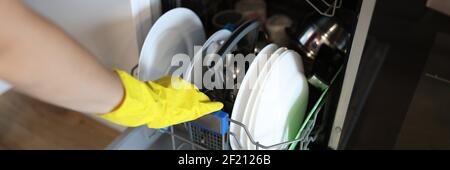 Mit der Hand der Frau in einem Gummihandschuh wird das Geschirr in die Spülmaschine gegeben Stockfoto