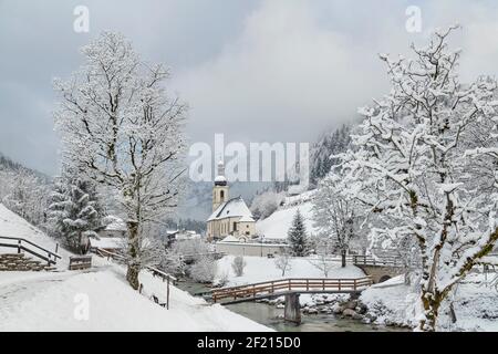 Deutschland, Bayern, Ramsau Dorf, berühmte aussicht auf die Kirche St. Sebastian mit der Fußgängerbrücke über die Ramsauer Ache in einer schneebedeckten Landschaft. Stockfoto