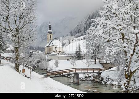 Deutschland, Bayern, Ramsau Dorf, berühmte aussicht auf die Kirche St. Sebastian mit der Fußgängerbrücke über die Ramsauer Ache in einer schneebedeckten Landschaft. Stockfoto