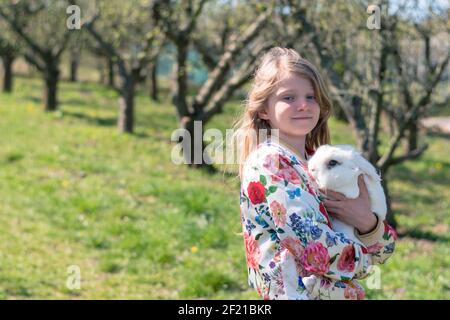 Kleines Mädchen, das im Gras sitzt und ein Weiß umarmt Flauschiges Kaninchen Haustier Stockfoto