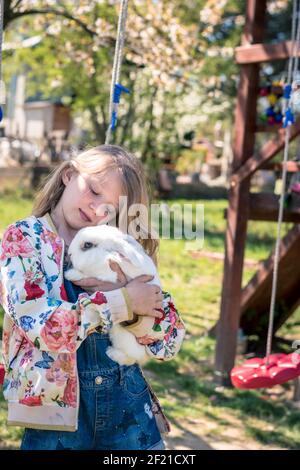 Schöne Teenager mit wilden lockigen langen blonden Haaren umarmt Weißes Kaninchen beim Sitzen im Garten Stockfoto