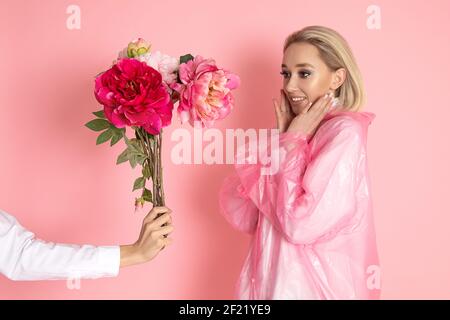 Hand in weißem Hemd gibt Bouquet von Pfingstrosen zu jungen blonden Frau auf rosa Hintergrund im Studio. Frau ist glücklich, Emotionen der Freude. Blumenlieferung Konz Stockfoto