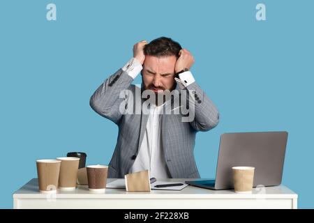 Junger Firmenarbeiter jammerte oder weinte, packte verzweifelt seinen Kopf und saß am Schreibtisch auf blauem Studiohintergrund Stockfoto