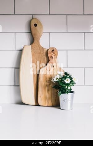 Weiße Gänseblümchen in einem silbernen Pflanzentopf neben zwei Holzhackbretter, die in einer Küche an weißen Fliesen gelehnt sind Stockfoto