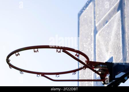 Ein Porträt eines Outdoor-Basketball-Felge ohne Netz und eine gefrorene Plexiglas-Backboard mit blauen Linien auf sie in immer durch Sonnenlicht beleuchtet. Die Basketba Stockfoto